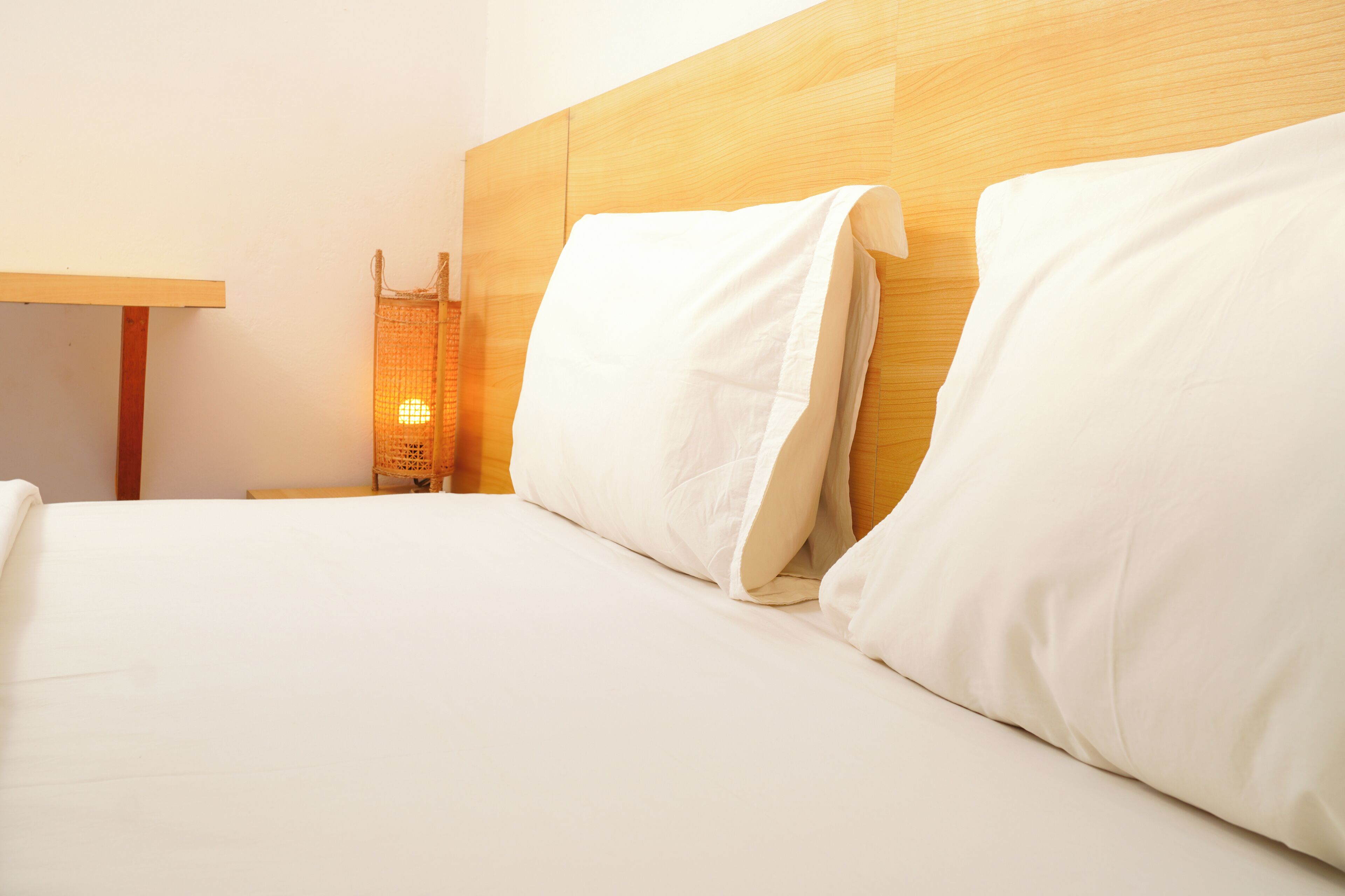 Una cama bien hecha con sábanas y almohadas blancas, un cabecero de madera y una mesita de noche con una lámpara.