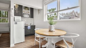 Une cuisine moderne avec des meubles blancs, un four à micro-ondes et un réfrigérateur. On y trouve une table à manger avec deux chaises, ainsi qu’un vase orné d’une plante.