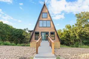 A triangular wooden cabin with a central door and windows, a wooden staircase leading up to the entrance, and a gravel path in front. - The Constellations - Romantic Cabins (Omaha)