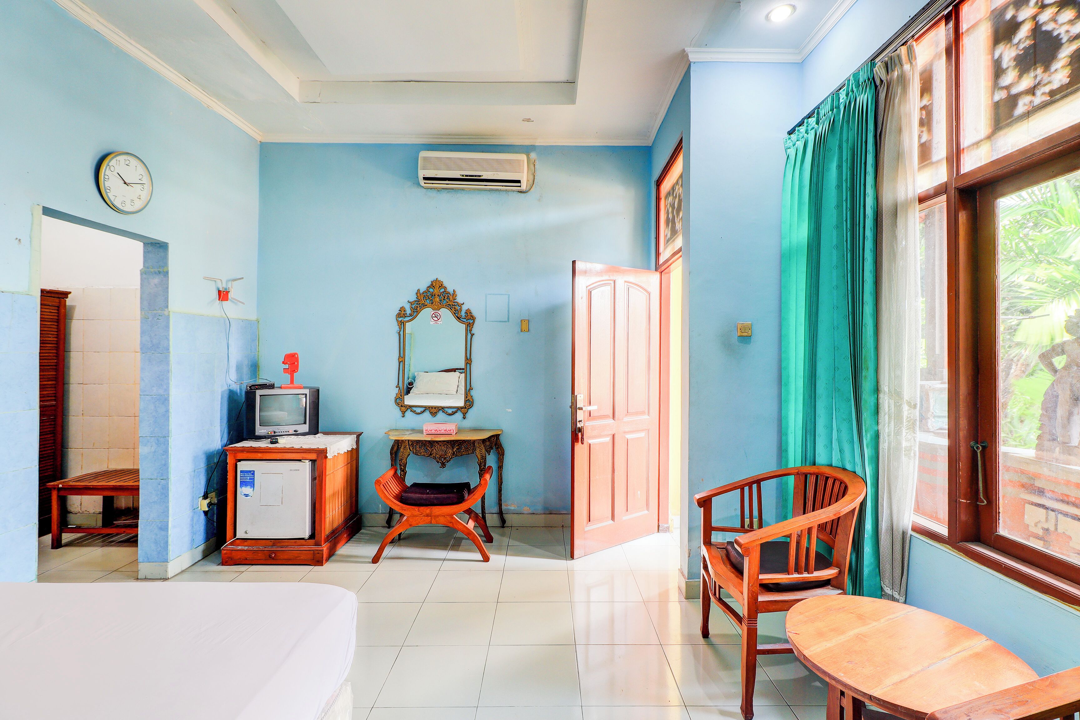 A room with a bed, a television, a small table, a chair, a mirror, and a window with curtains.
