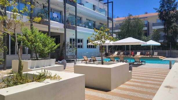 A modern outdoor pool area with lounge chairs and umbrellas, surrounded by planters and a building with balconies. - Mythical Sands- HANA (Protaras)