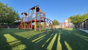 A playground with a climbing structure and slides, surrounded by a fenced area and trees. - Mythical Sands- HANA (Protaras)