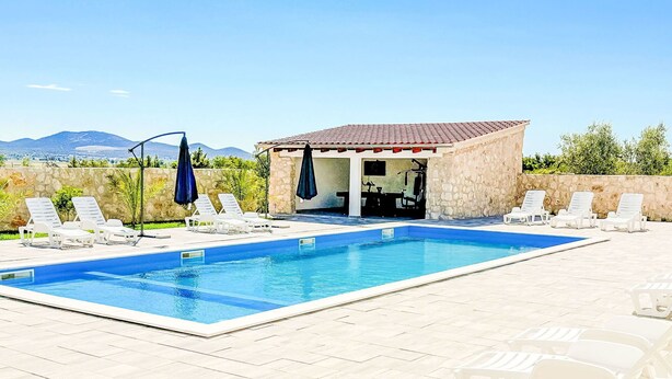 Una piscina rectangular con sillones de descanso blancos y sombrillas, un área techada con equipo de ejercicio y un muro de piedra.