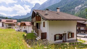 A two-story chalet with a sloped roof, wooden balconies, and a view of mountains in the background.