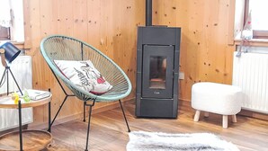 A cozy room with a wooden floor, a wicker chair with a cushion, a small round table with a lamp, a white fluffy ottoman, and a black wood-burning stove.