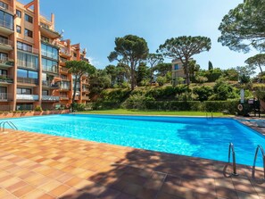 Piscina com deck ladrilhado, rodeada por um edifício de vários andares com varandas e um morro com árvores.