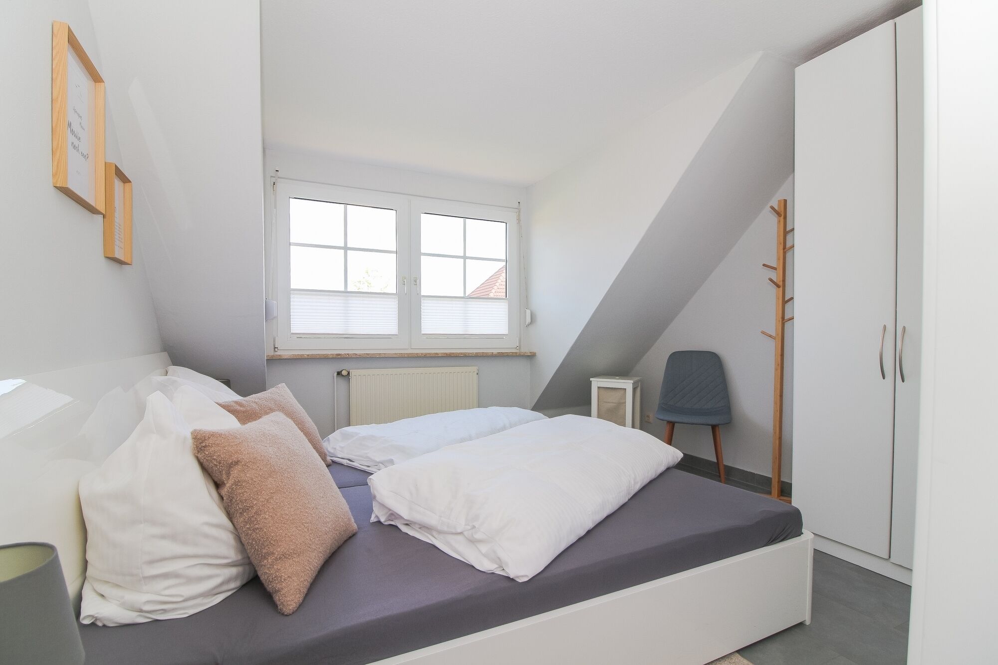 Une chambre avec un plafond en pente, un lit avec une literie blanche et grise, une fenêtre avec des stores, un radiateur, une chaise et une armoire.