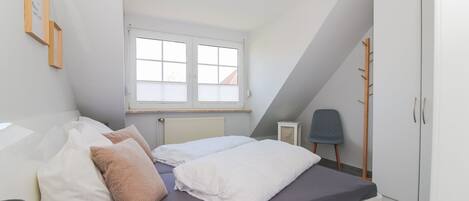 Un dormitorio con techo inclinado, cama con ropa de cama blanca y gris, ventana con persianas, radiador, una silla y un armario.