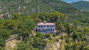 A two-story house with a balcony, surrounded by dense greenery and hills in the background.