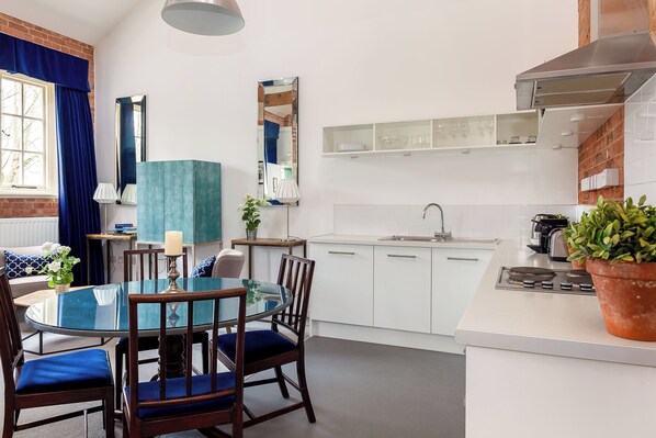 Una cocina moderna con armarios blancos, estufa integrada y una zona de comedor azul con mesa de vidrio y sillas de madera.