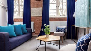 A cozy living room with a sofa, a coffee table, and a chair. There is a window with blue curtains and a view of trees. - Bowen-Buscarlet at Bicester Heritage (Bicester)