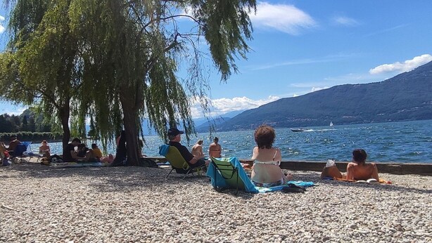 A pebbled beach with people relaxing under a large tree, overlooking a lake with boats and mountains in the background. - Nice apartment for 4 people with A/C, WIFI, TV and balcony (Porto Valtravaglia)