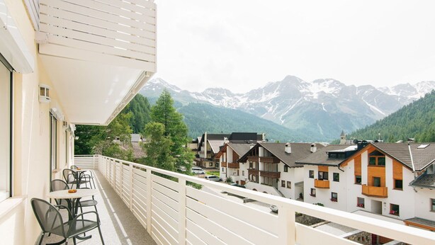 A balcony with a table and chairs, overlooking a mountain range and a row of houses.