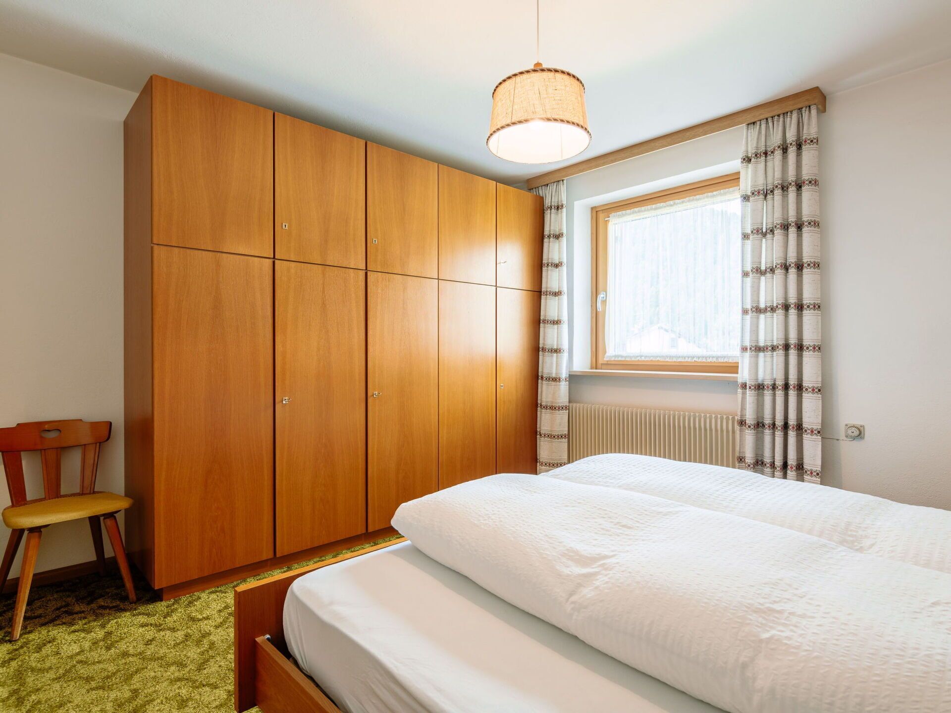 A bedroom with a wooden wardrobe, a single bed, a chair, and a window with curtains.