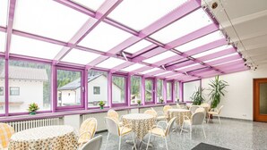 A sunlit room with a glass ceiling, multiple tables and chairs, and a view of houses outside.