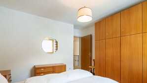 A bedroom with a bed, a wooden wardrobe, a round mirror, and a hanging light fixture.