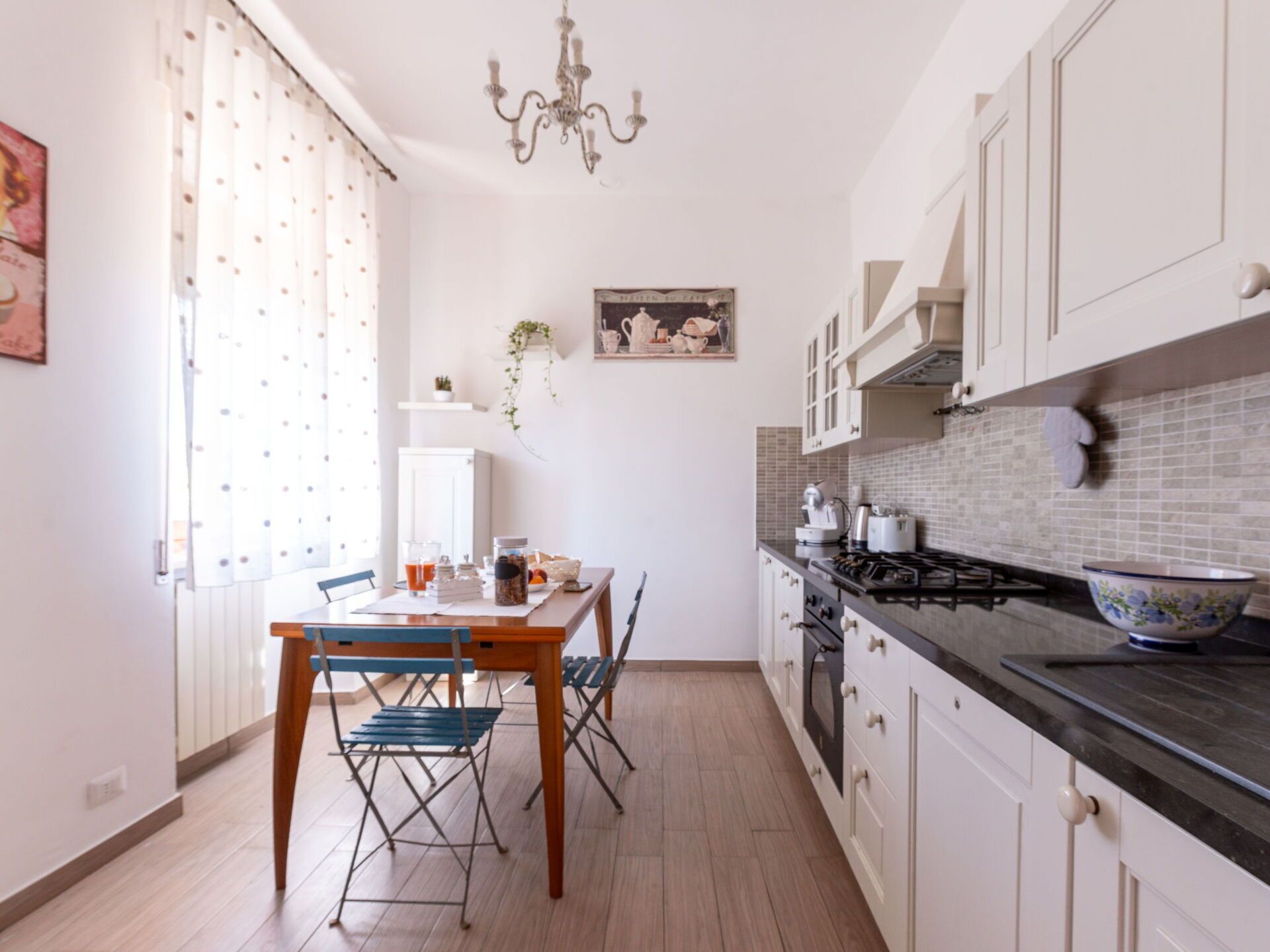 A kitchen with white cabinets, a black countertop, and a chandelier. There is a dining table with chairs and a window with curtains.