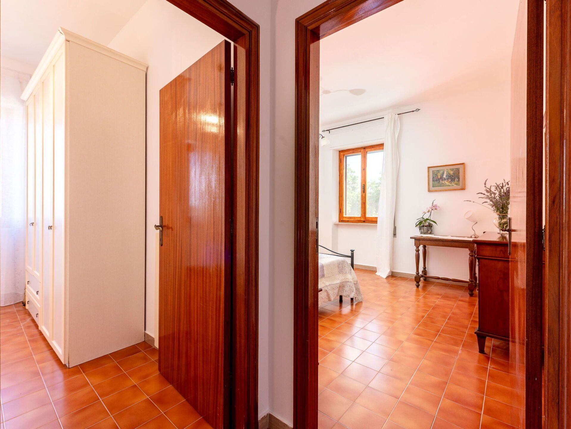 Un couloir avec des portes en bois, un sol carrelé, une fenêtre avec des rideaux, un bureau avec une lampe et un tableau accroché au mur.