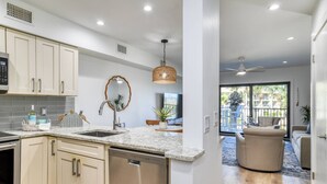A modern kitchen with white cabinets, a granite countertop, and a dishwasher. There is a dining area with a table and chairs, and a view of a balcony with outdoor furniture.