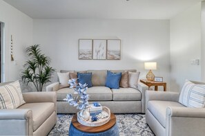 Ett vardagsrum med en beige soffa, två fåtöljer, ett soffbord med dekorativa föremål, ett sidobord med en lampa och konst på väggen.