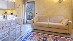 Een woonkamer met een beige bank, een vloerkleed met een patroon, een eettafel met stoelen, een keukenhoek en een balkon met uitzicht op de heuvels.
