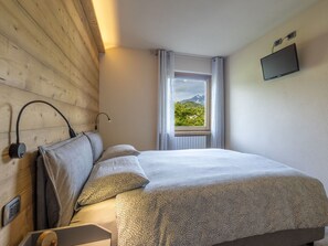 Een slaapkamer met een houten hoofdeinde, een bed met beddengoed met een patroon, een raam met uitzicht op groen en bergen, en een flatscreen televisie aan de muur.