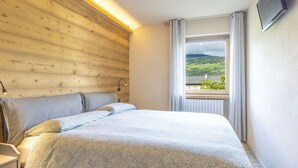 Een slaapkamer met een houten hoofdbord, een bed met beddengoed met een patroon, een nachtkastje, een bedlampje, een raam met uitzicht op groen en een aan de muur bevestigde televisie.
