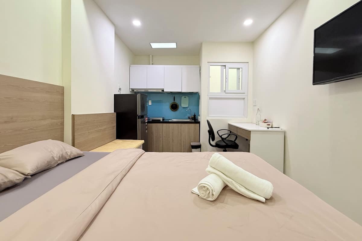 A compact hotel room with a bed, a desk, a chair, a small kitchenette, and a TV mounted on the wall.