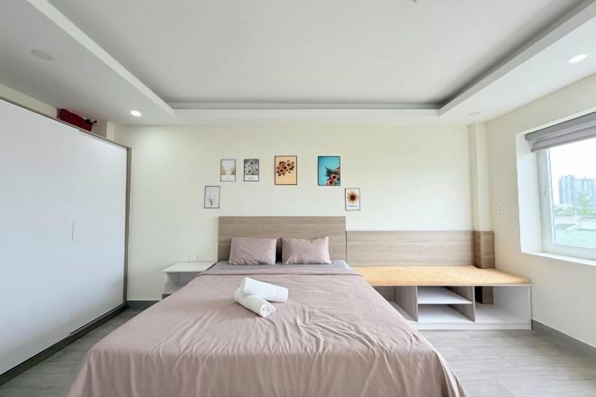 A modern hotel room with a large bed, a bedside table, a bench at the foot of the bed, and a window with a city view.
