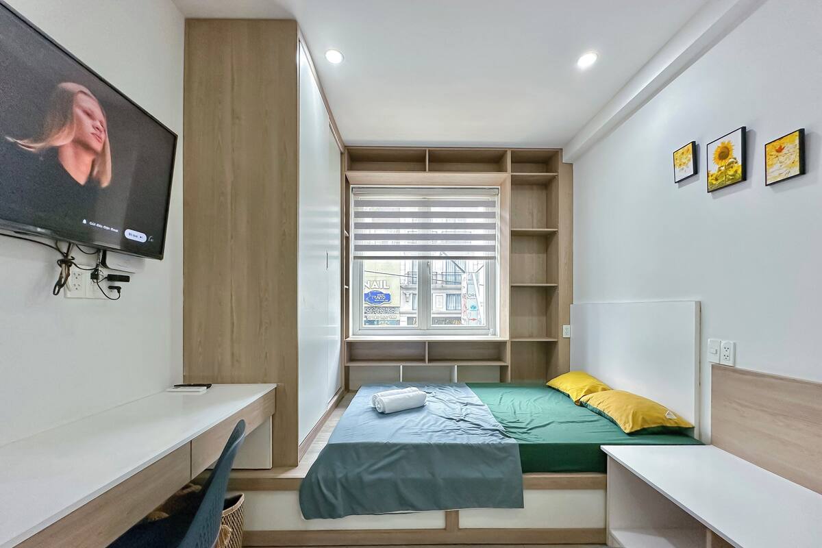A compact bedroom with a bed, a desk, a TV mounted on the wall, and a window with blinds.