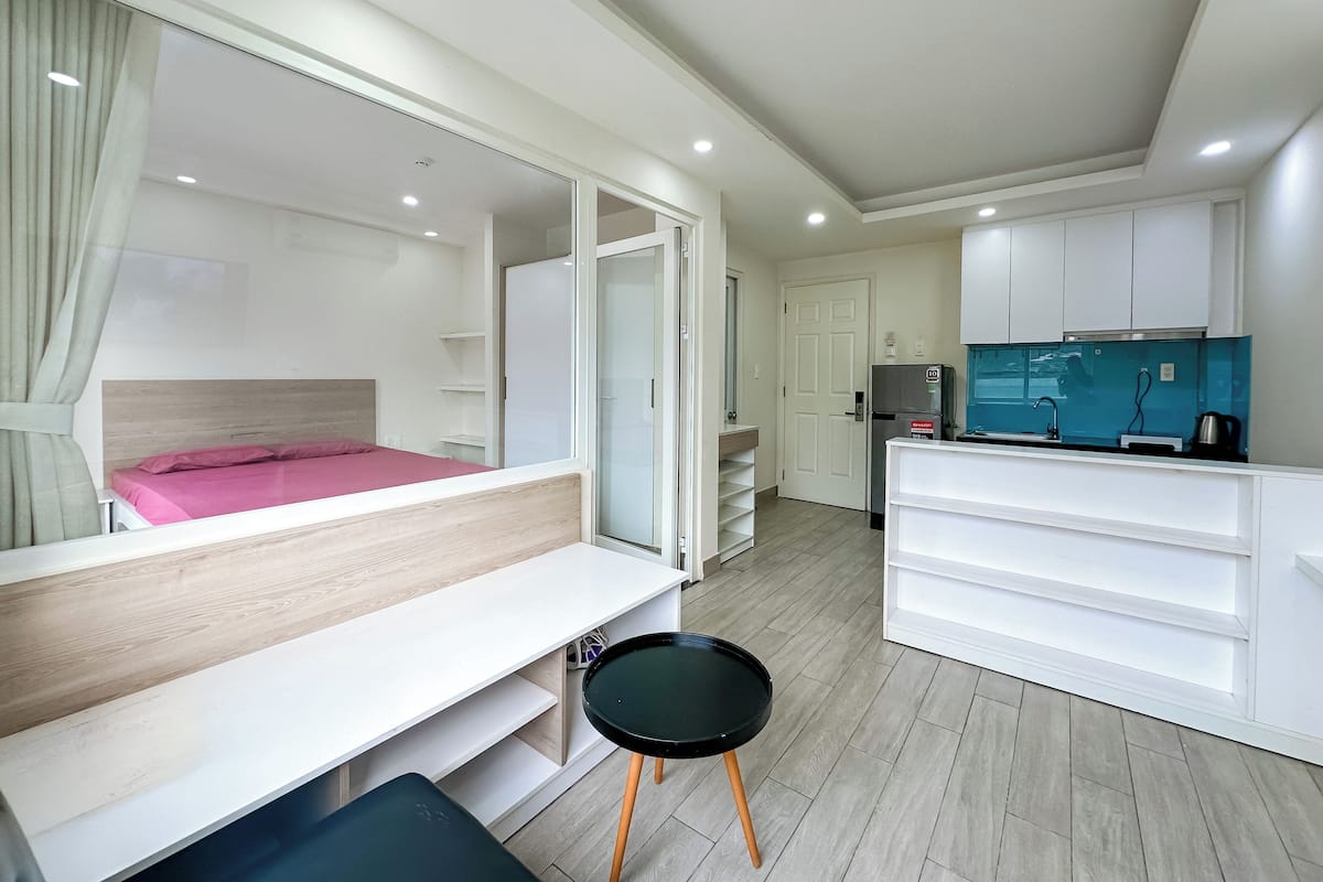 A modern hotel room with a bed, a desk, a chair, a small table, a kitchenette, and a bathroom.