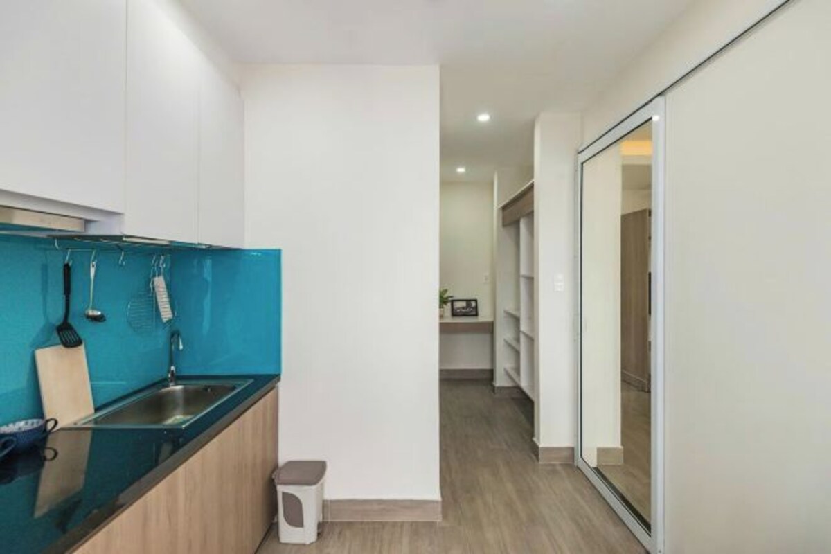A narrow hallway with a kitchen area on the left, featuring a sink, countertop, and hanging utensils. A small stool is placed near the sink. The hallway leads to a room with a sliding glass door on the right.