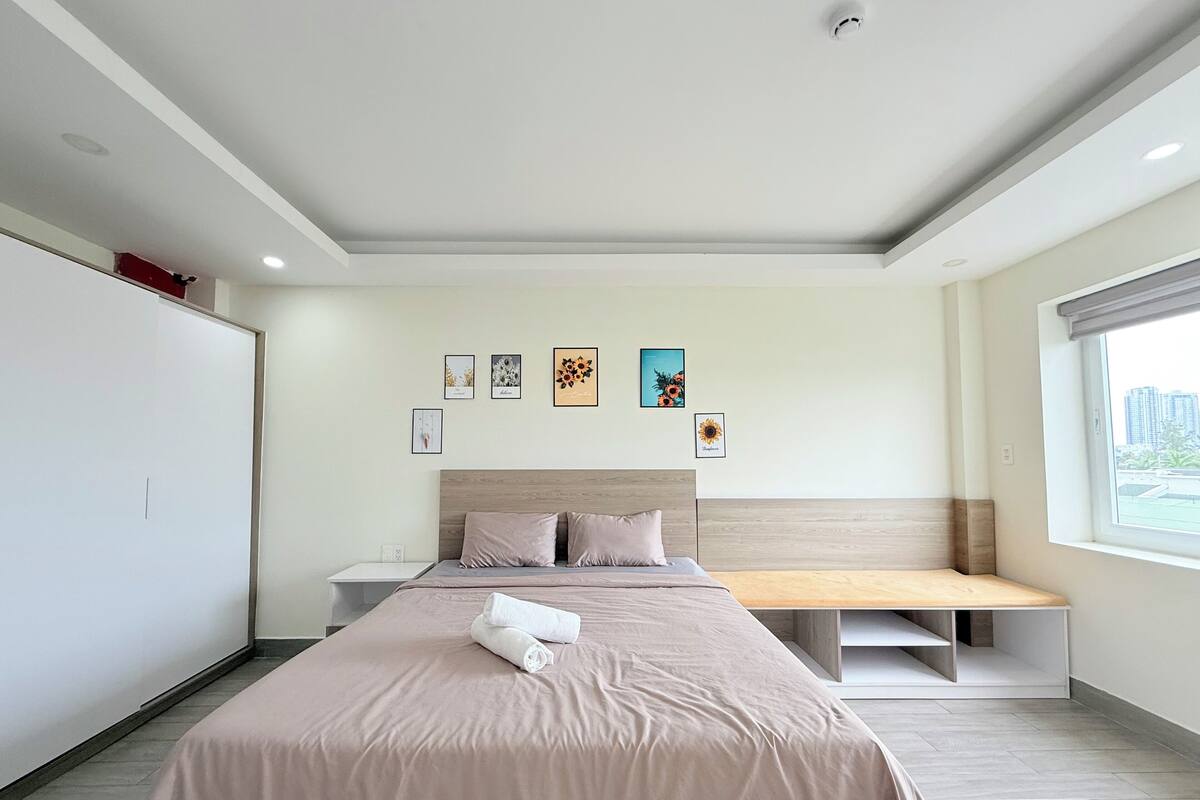 A modern hotel room with a large bed, a bedside table, a bench at the foot of the bed, and a window with a city view.