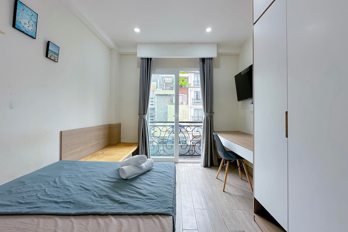 A bedroom with a bed, a desk, a TV, and a balcony with a view of buildings.