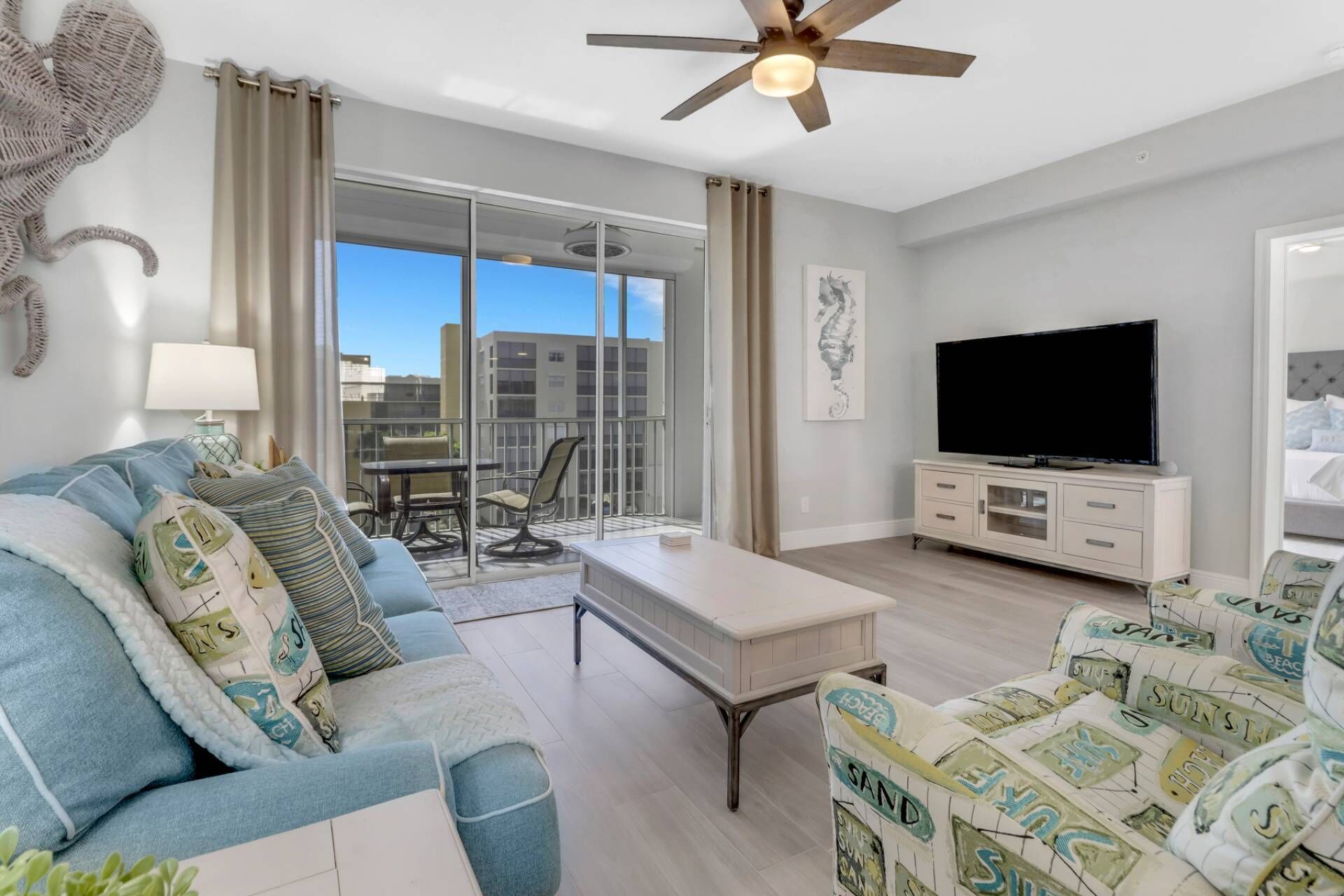 A modern living room with a sofa, a coffee table, a TV on a cabinet, and a balcony with a view of buildings.