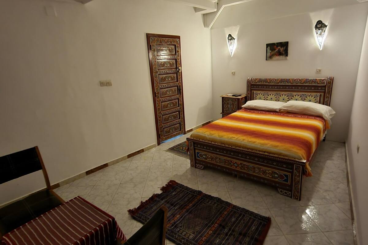 A bedroom with a bed, a chair, a door, and a wall-mounted light fixture.