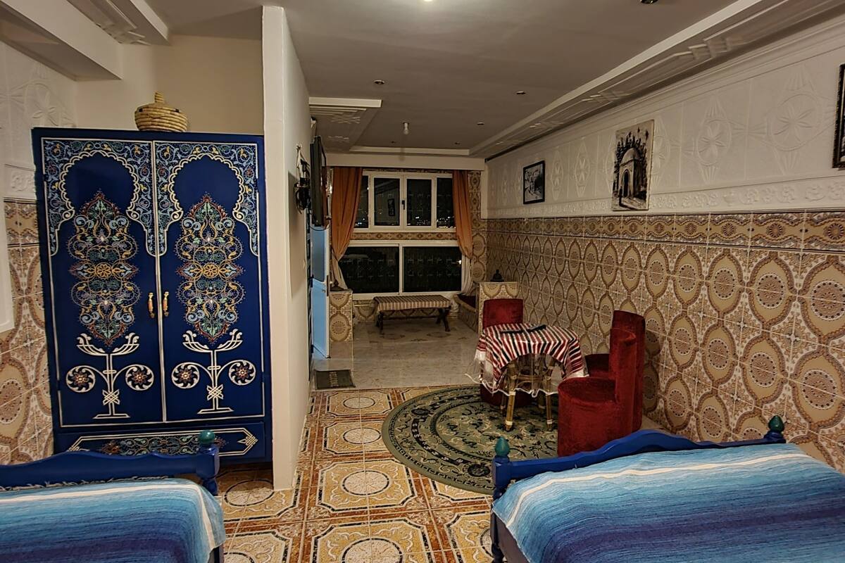 A hotel room with two beds, a sitting area with a red chair, a small table, and a decorative cabinet.