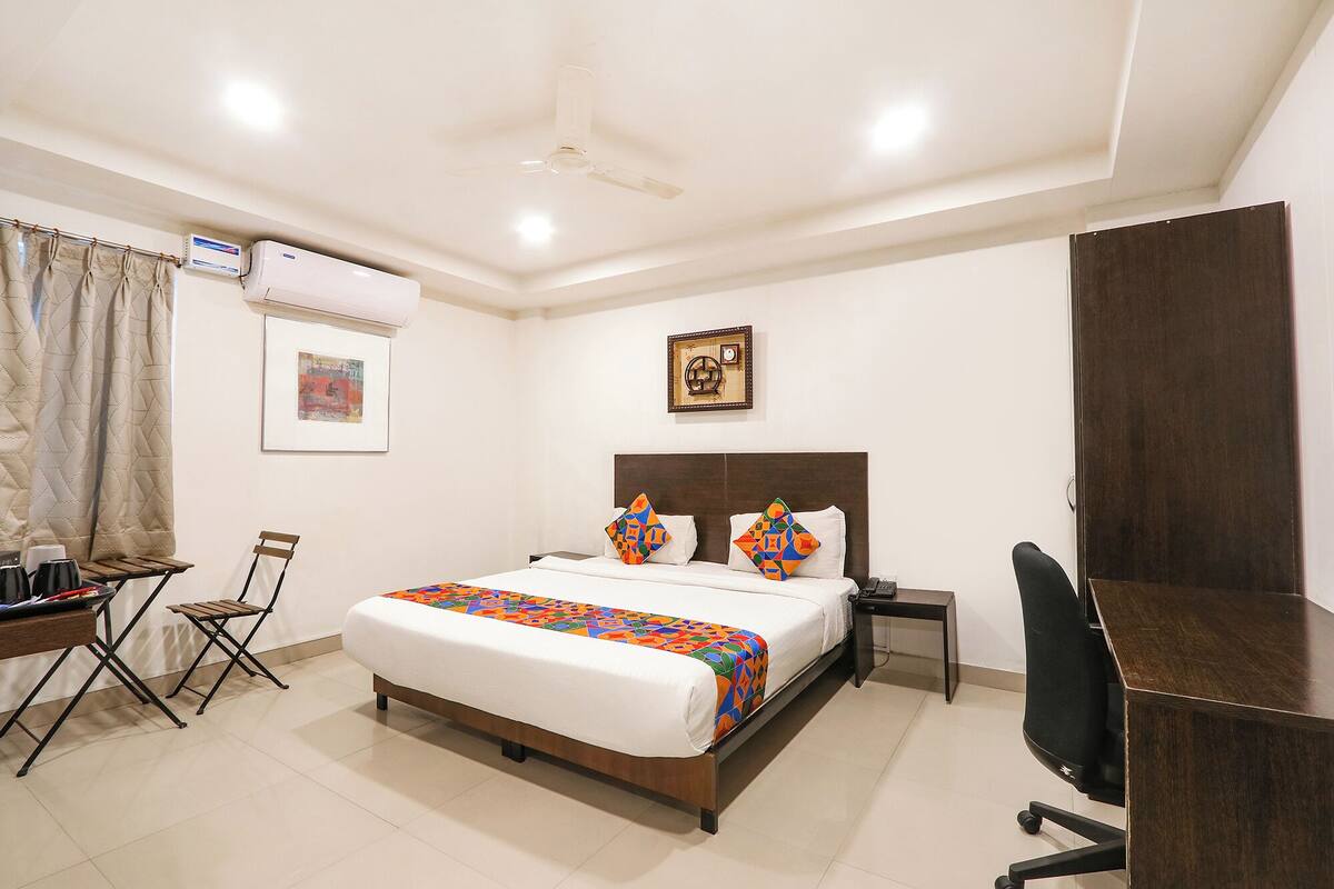 A hotel room with a bed, desk, chair, and an air conditioning unit.