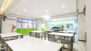 Un espace de restauration moderne avec des tables et des chaises blanches, de grandes fenêtres donnant sur un paysage verdoyant et un ventilateur de plafond.