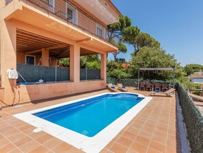 Ein Schwimmbad mit überdachter Terrasse, Gartenmöbeln und Blick auf Bäume sowie ein Gebäude im Hintergrund.