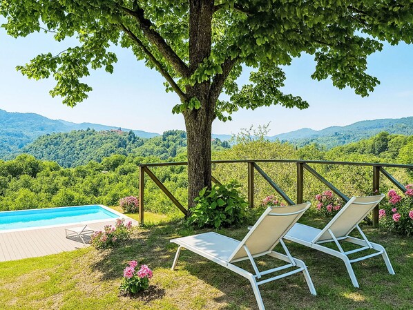 A pool with two lounge chairs under a tree, overlooking a mountainous landscape.