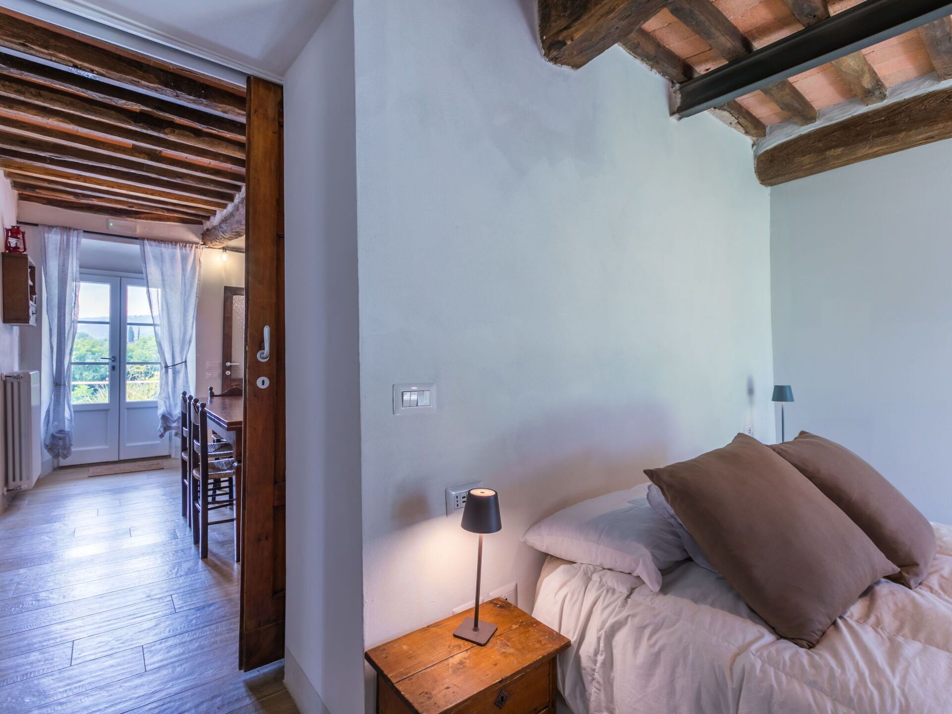 A bedroom with a wooden nightstand, a lamp, a bed with pillows, and a view of a balcony with a door leading outside.