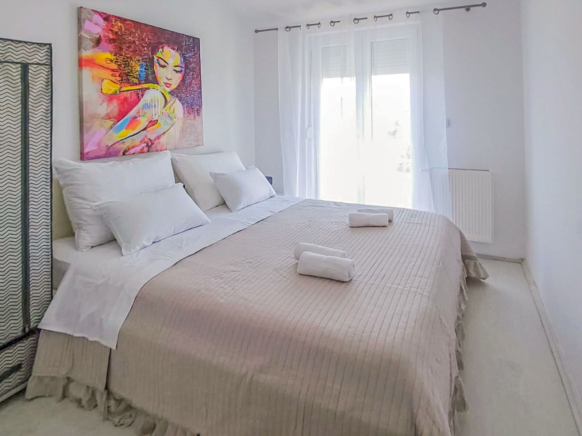 Un dormitorio con una cama grande, ropa de cama blanca, un cuadro colorido sobre la cama y una ventana con cortinas.
