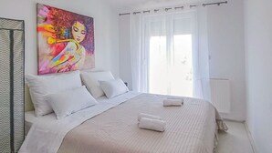 Une chambre à coucher avec un grand lit, du linge de lit blanc, un tableau coloré au-dessus du lit et une fenêtre avec des rideaux.