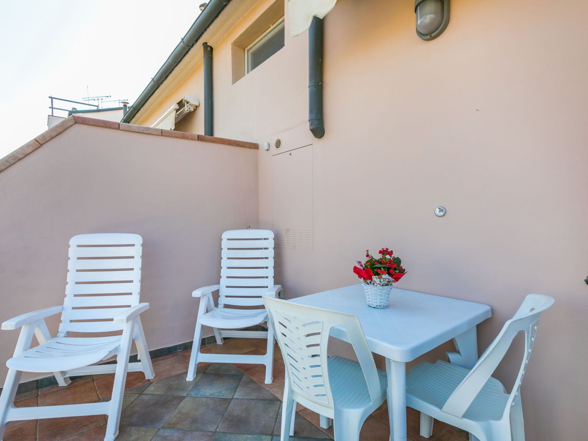 An outdoor patio with white plastic chairs and a table, a small potted plant, and a wall-mounted light fixture.