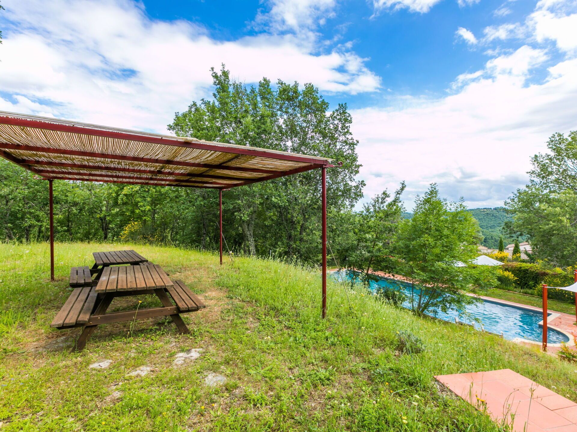 Un área de picnic con una mesa y bancos de madera, un espacio de estar cubierto y una piscina.