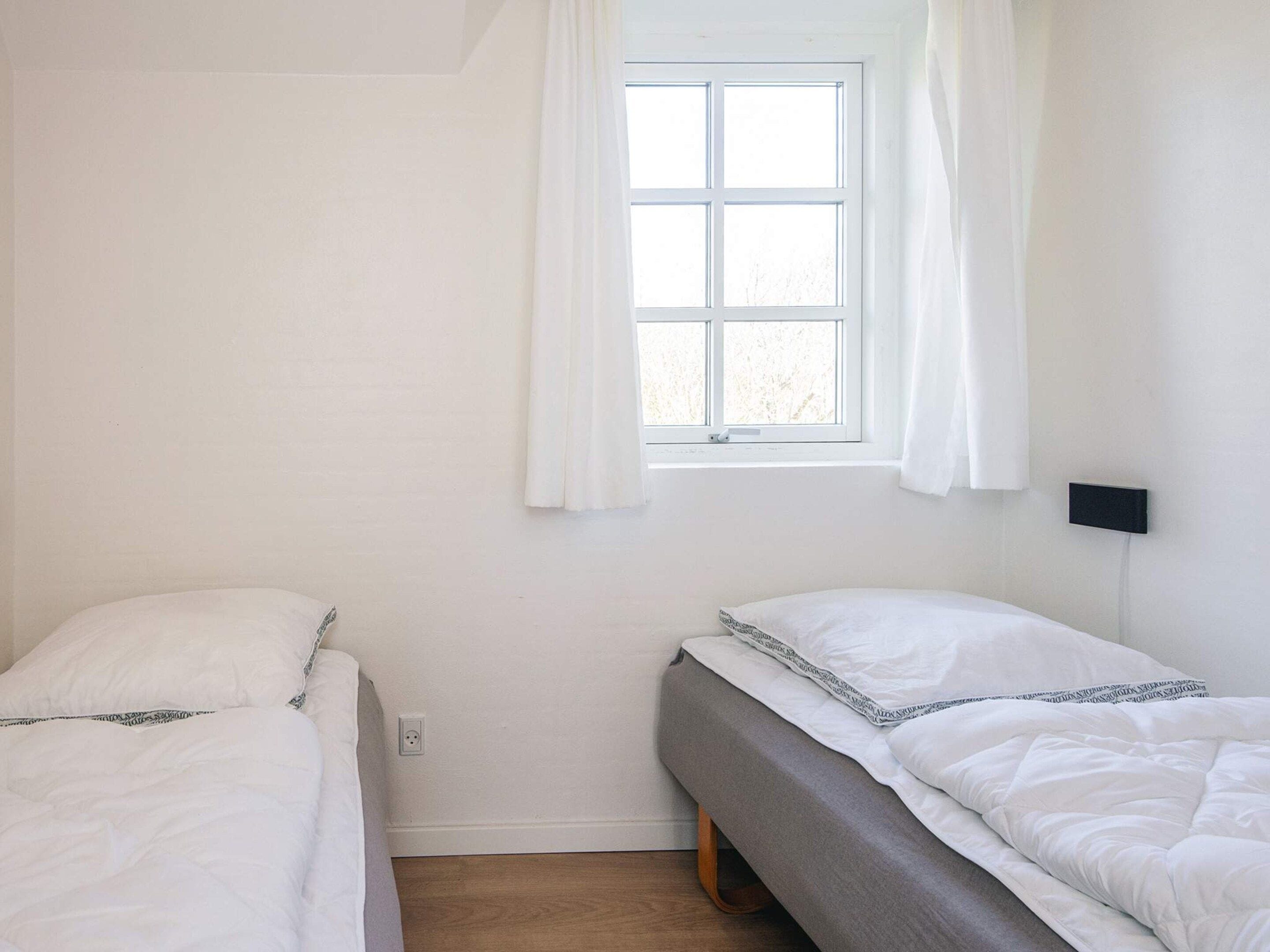 A room with two beds, a window with curtains, a wall socket, and a black switchboard.