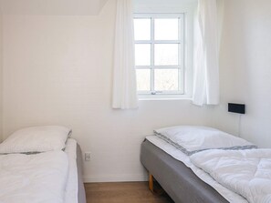 Une chambre avec deux lits simples, une fenêtre avec des rideaux blancs, un luminaire fixé au mur et un plancher en bois.