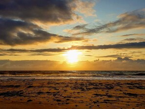 Ein Strand mit untergehendem Sonnenuntergang am Horizont, Wolken am Himmel und ansteigenden Wellen.