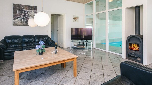Ein modernes Wohnzimmer mit Holzbalkendecke, einem schwarzen Ledersofa, einem Couchtisch aus Holz mit einer Vase und Gläsern, einem Flachbildfernseher und einem Kaminofen.